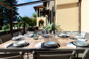 Un área de comedor al aire libre con mesas dispuestas para una comida, rodeada de vegetación y un edificio al fondo.