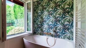 Une salle de bain avec une baignoire sur pieds, une fenêtre donnant sur un paysage verdoyant et un mur orné d’un motif de feuilles.