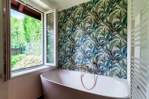 Un baño con una bañera independiente, un ventanal con vistas a zonas verdes y una pared con estampado de hojas.