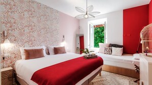 Une chambre avec un grand lit, un mur d’accent rouge, une fenêtre donnant sur un paysage verdoyant et une cage à oiseaux avec un oiseau à l’intérieur.