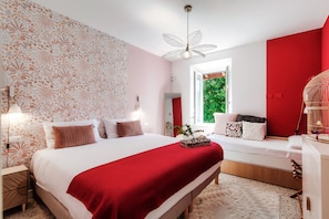 Un dormitorio con una cama grande, una pared de acento roja, un ventanal con vistas a la vegetación y una jaula de pájaros con un ave.