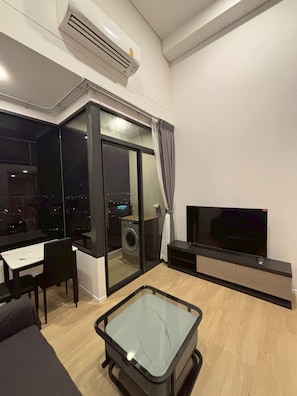 Uma sala de estar moderna com uma mesa de centro de vidro, televisão de tela plana, máquina de lavar roupa e um espaço de jantar com mesa e cadeiras.