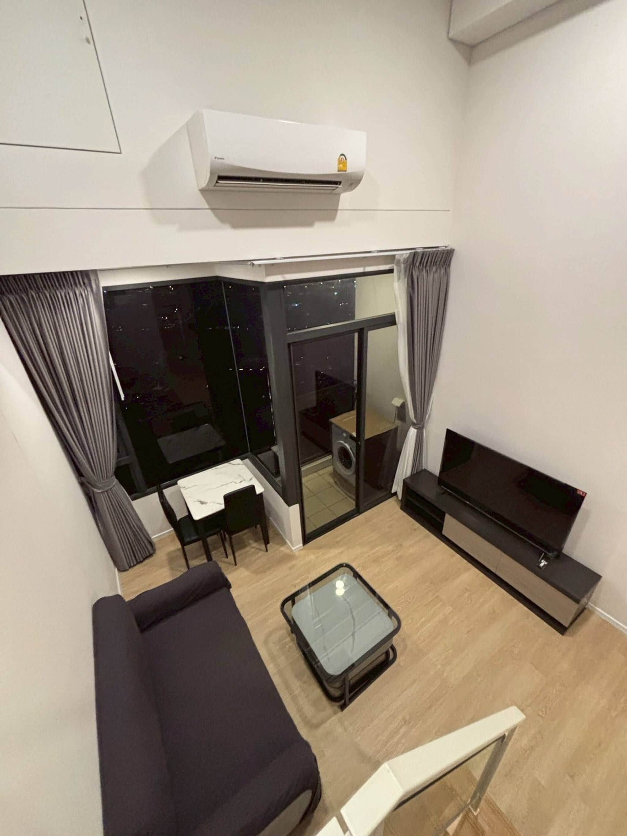 A modern living room with a sofa, a glass coffee table, a flat-screen TV, and a sliding glass door leading to a balcony.
