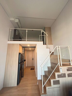 Un interior moderno con una escalera que sube a un altillo, una cocina con refrigerador y una puerta que da a otra habitación.