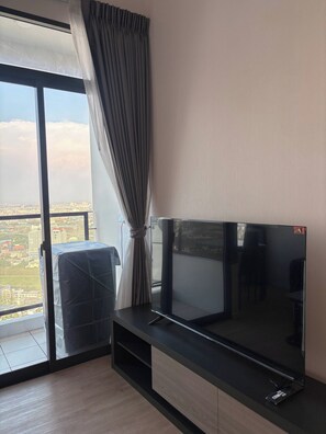 Una sala de estar moderna con un televisor de pantalla plana grande sobre un mueble de madera, un puerta corrediza de vidrio que da a un balcón con vista a la ciudad, y una cortina medio bajada.