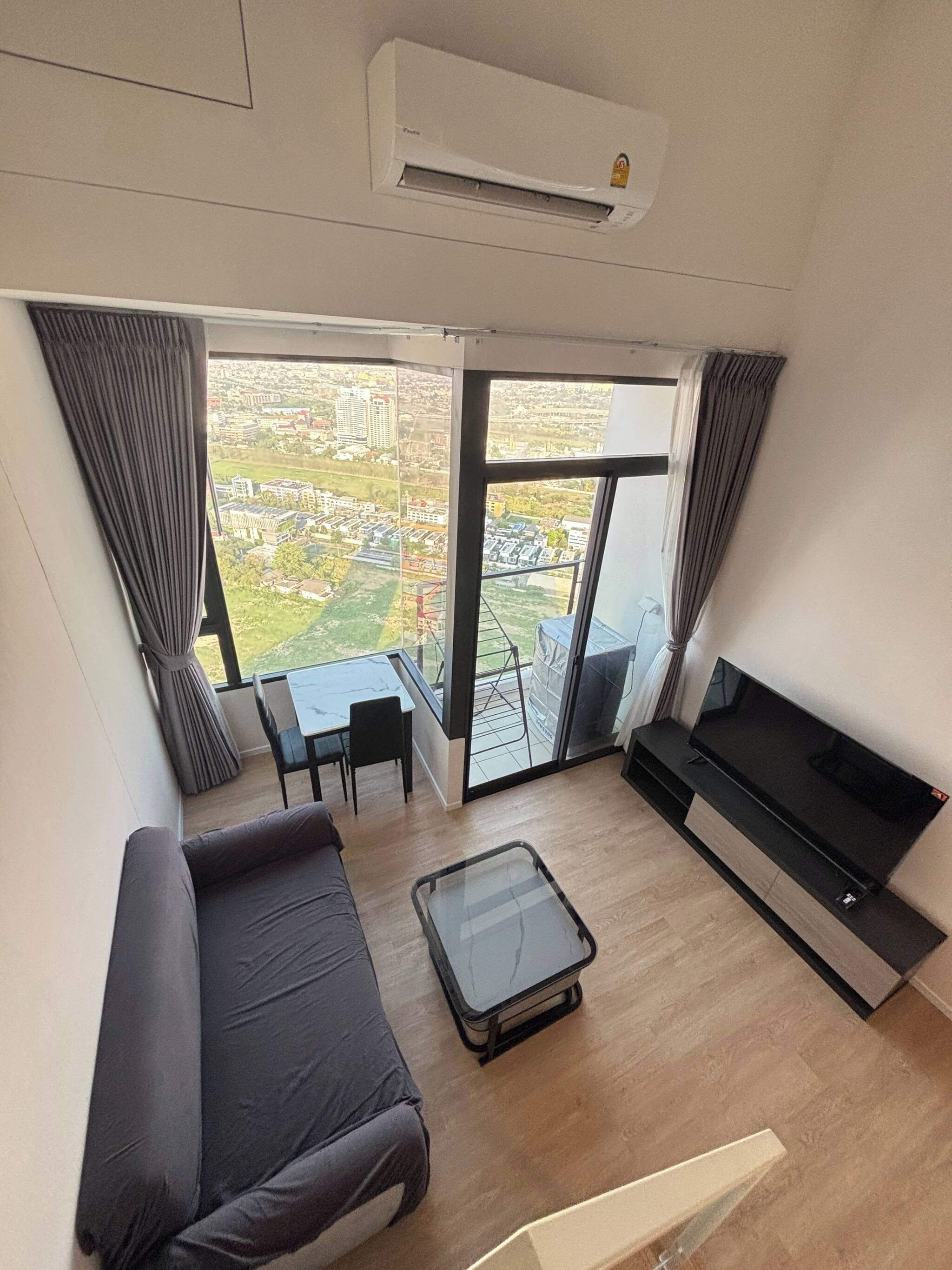 A modern living room with a sofa, a TV, a glass table, and a balcony with a view of a cityscape.