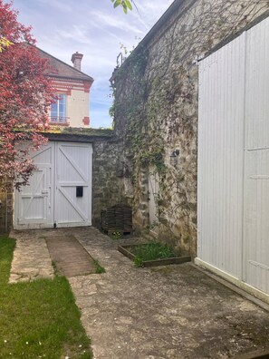 A courtyard with a white garage door, a stone wall with climbing plants, and a small garden area. - Charming country house (Villiers-sous-Grez)