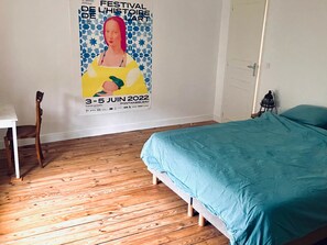 A bedroom with a wooden floor, a bed with a teal bedspread, a white chair, and a poster on the wall advertising a festival. - Charming country house (Villiers-sous-Grez)