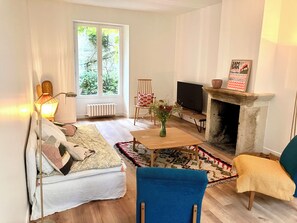 A living room with a sofa, a blue chair, a fireplace, a television, and a window with a view of greenery. - Charming country house (Villiers-sous-Grez)