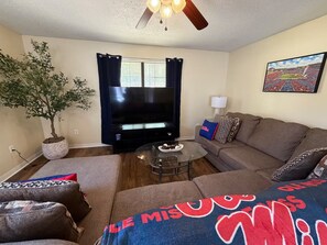 A living room with a flat-screen TV, a ceiling fan, a potted plant, and a couch with patterned cushions. - 6 min drive to Ole Miss & Square--Pet Friendly (Oxford)