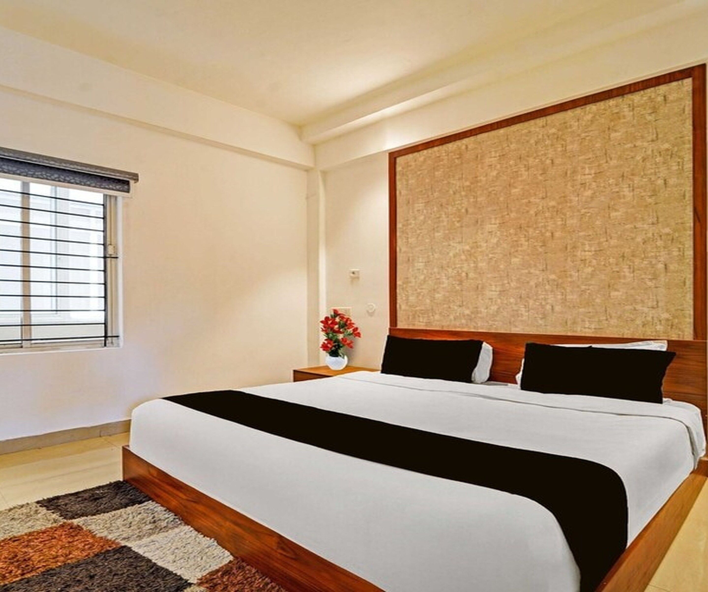 A hotel room with a large bed, a window with blinds, a nightstand with a vase of flowers, and a textured wall panel.