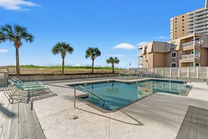Ein Schwimmbad mit Liegestühlen, einer Holzterrasse und Blick auf Strand und Gebäude.