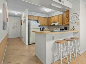 A kitchen with wooden cabinets, a refrigerator, a clock, and a bar area with stools. - 3BR Pelicans Watch 110- Pool- 3BR In Shore Drive (Myrtle Beach)