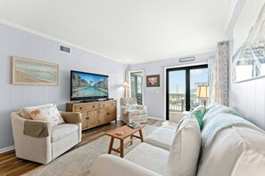 Ein Wohnzimmer mit Fernseher, einer Couch, einem Sessel und einem kleinen Tisch. Eine Tür führt auf einen Balkon.