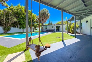 A covered outdoor area with a pool, artificial grass, and a swing set. - Luxury 6BR 2-Home Retreat w/ Pool, BBQ & Games (Pompano Beach)