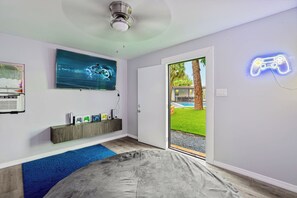 A modern bedroom with a bed, a ceiling fan, a wall-mounted TV, and a neon game controller sign. - Luxury 6BR 2-Home Retreat w/ Pool, BBQ & Games (Pompano Beach)