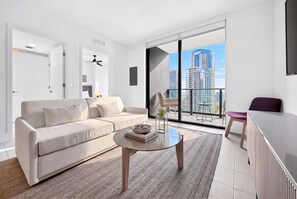 A modern living room with a sofa, a coffee table, and a view of high-rise buildings. - Luxury 1BR King Condo with Balcony, Pool & Spa (Miami)