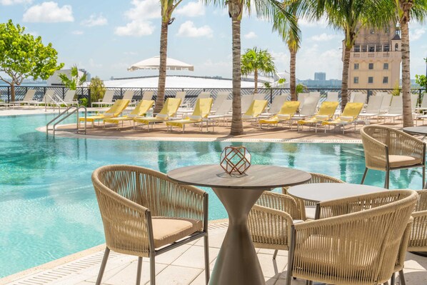 A poolside area with lounge chairs and a table, surrounded by palm trees and a building in the background. - Luxury 1BR King Condo with Balcony, Pool & Spa (Miami)