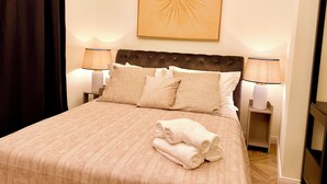Ein ordentlich bezogenes Bett mit einer beigen Tagesdecke, zwei weißen Kissen und einem gefalteten weißen Handtuch. Darüber hängt ein Wandbild und auf beiden Seiten des Bettes stehen Nachttischlampen.