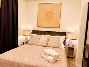 Una cama bien tendida con una colcha color beige, dos almohadas blancas y una toalla blanca doblada. Hay una obra de arte colgada en la pared sobre la cama y dos lámparas de noche a cada lado.