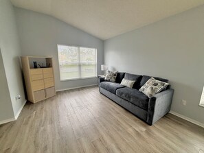 Ein Zimmer mit einem grauen Sofa, einem Holzschrank und einem Fenster mit Jalousien.