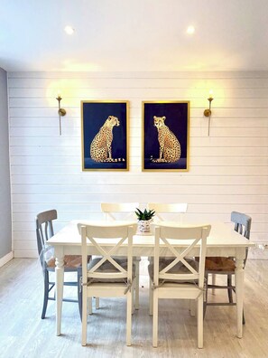 Ein Essbereich mit einem weißen Tisch und Stühlen, zwei gerahmte Gepardenbilder an der Wand und Wandleuchten.