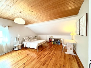 Ein Schlafzimmer mit Holzboden, einem Bett, einem Nachttisch, einem Sessel, einem Bücherregal und einem Fenster.