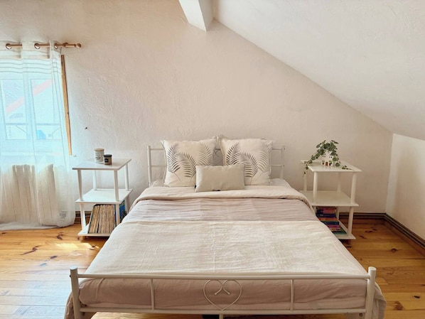 Ein Schlafzimmer mit einem Bett, zwei Nachttischen, einem Fenster mit Vorhängen und einem kleinen Tisch mit einer Pflanze.