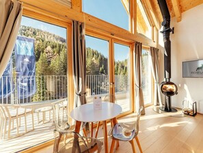 A room with a round table and chairs, large windows with curtains, a fireplace, and a view of a forested area. - Chalet # Rfl006 for 2 People With Sauna (Oberwölz)