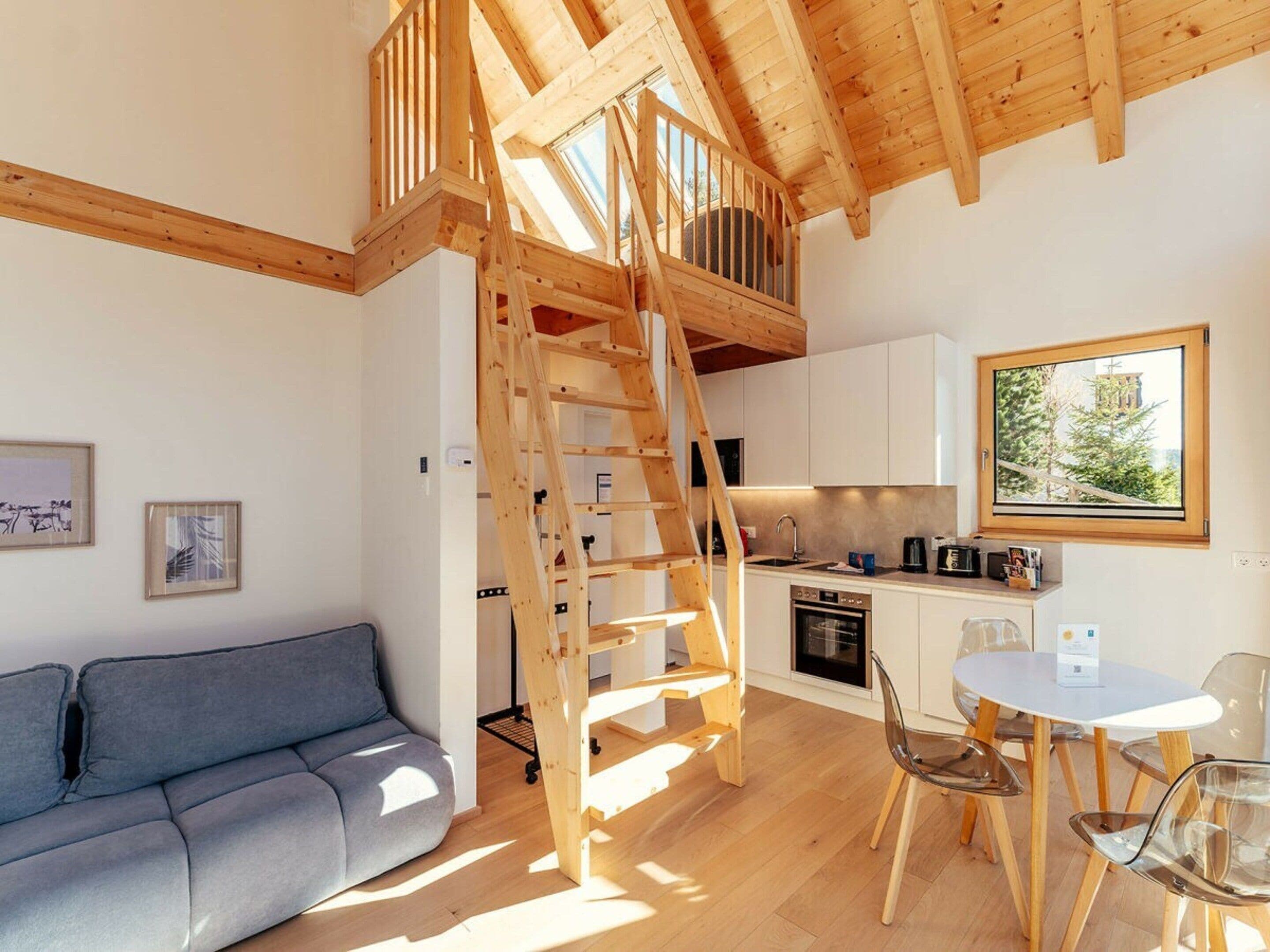 Ein modernes Interieur mit einer Holztreppe, die zu einer oberen Ebene führt, einem Küchenbereich und einer Sitzecke mit Sofa und Sesseln.