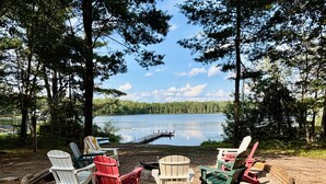 Ein Sitzbereich am Seeufer mit Adirondack-Stühlen, die um eine Feuerstelle angeordnet sind.
