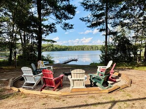 A lakeside seating area with Adirondack chairs arranged around a fire pit. - Cozy 2-bedroom house in tranquil Frederic with AC (Frederic)