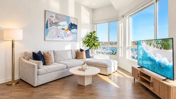 A modern living room with a sofa, coffee table, and TV, featuring a large window with a view of the sea.