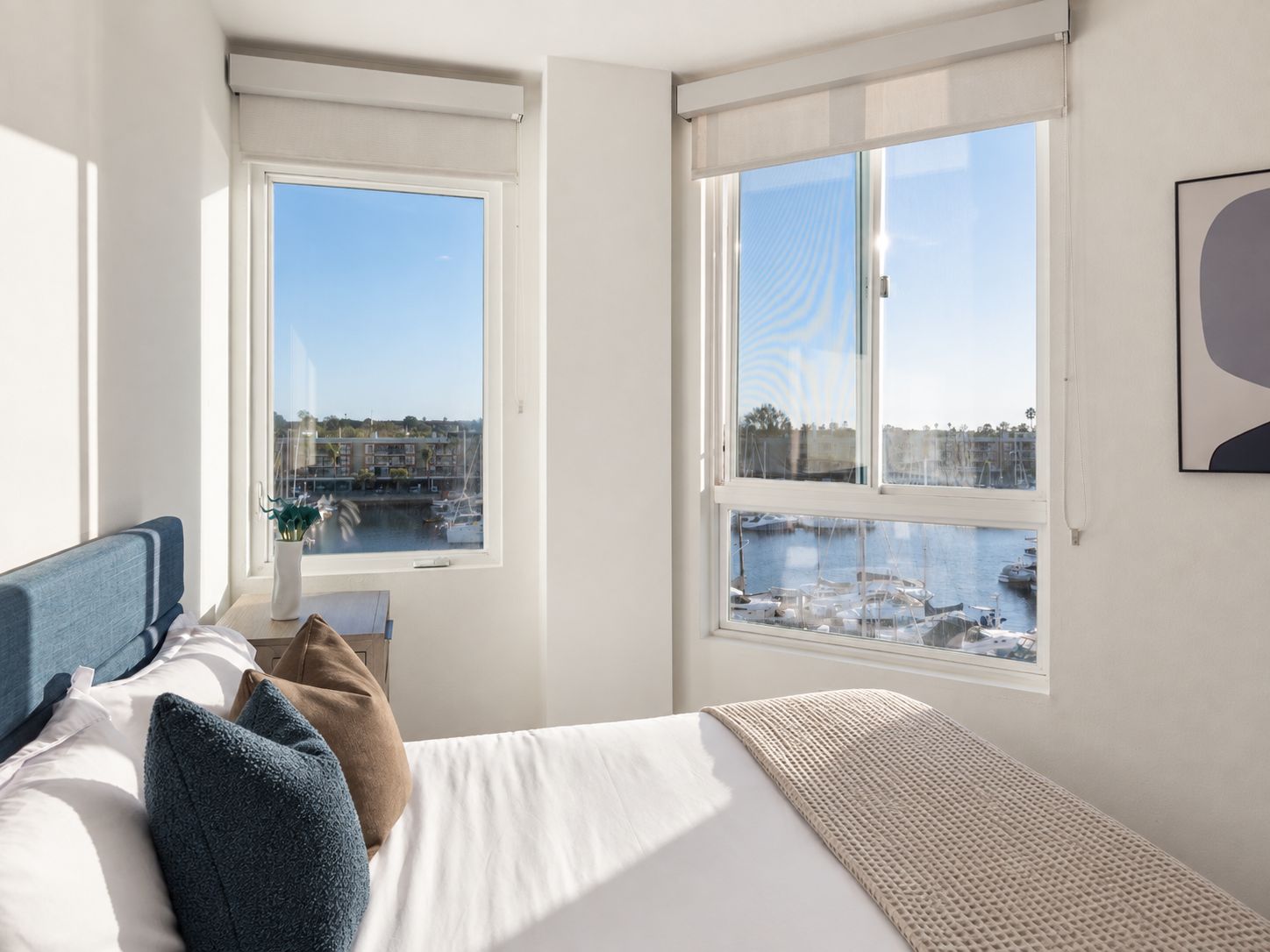 Una habitación de hotel con una cama, una ventana con vista a un puerto deportivo y un cuadro en la pared.