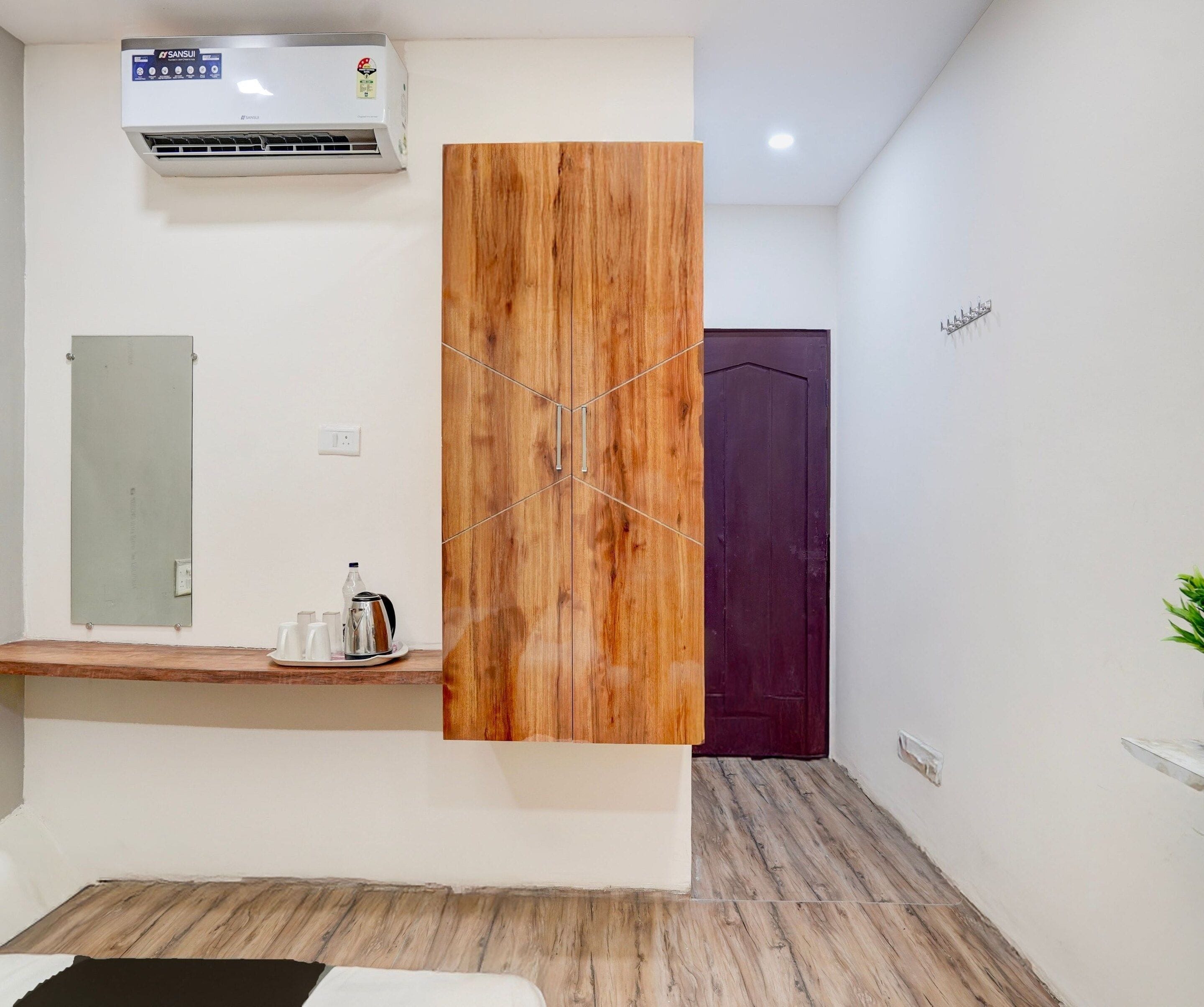 Habitación de hotel con armario de madera, un espejo, una mesita con hervidor y tazas, una puerta morada y un equipo de aire acondicionado.