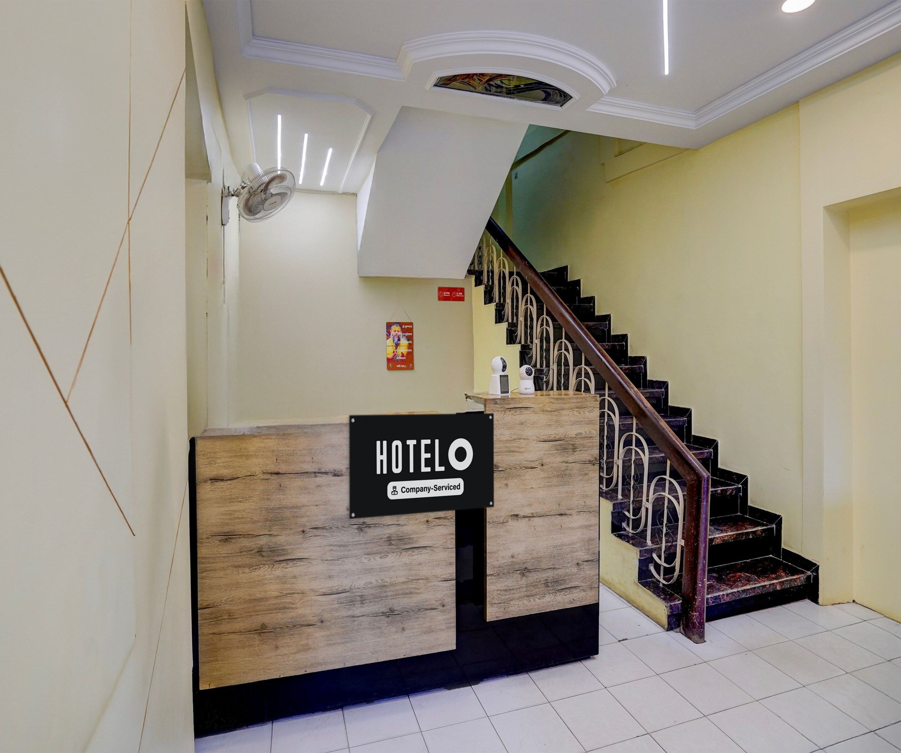 A hotel reception desk with a wooden finish and a sign indicating it is company-serviced. A staircase with a metal railing leads to an upper level.