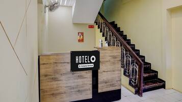 A hotel reception desk with a wooden finish and a sign indicating it is company-serviced. A staircase with a metal railing leads to an upper level.