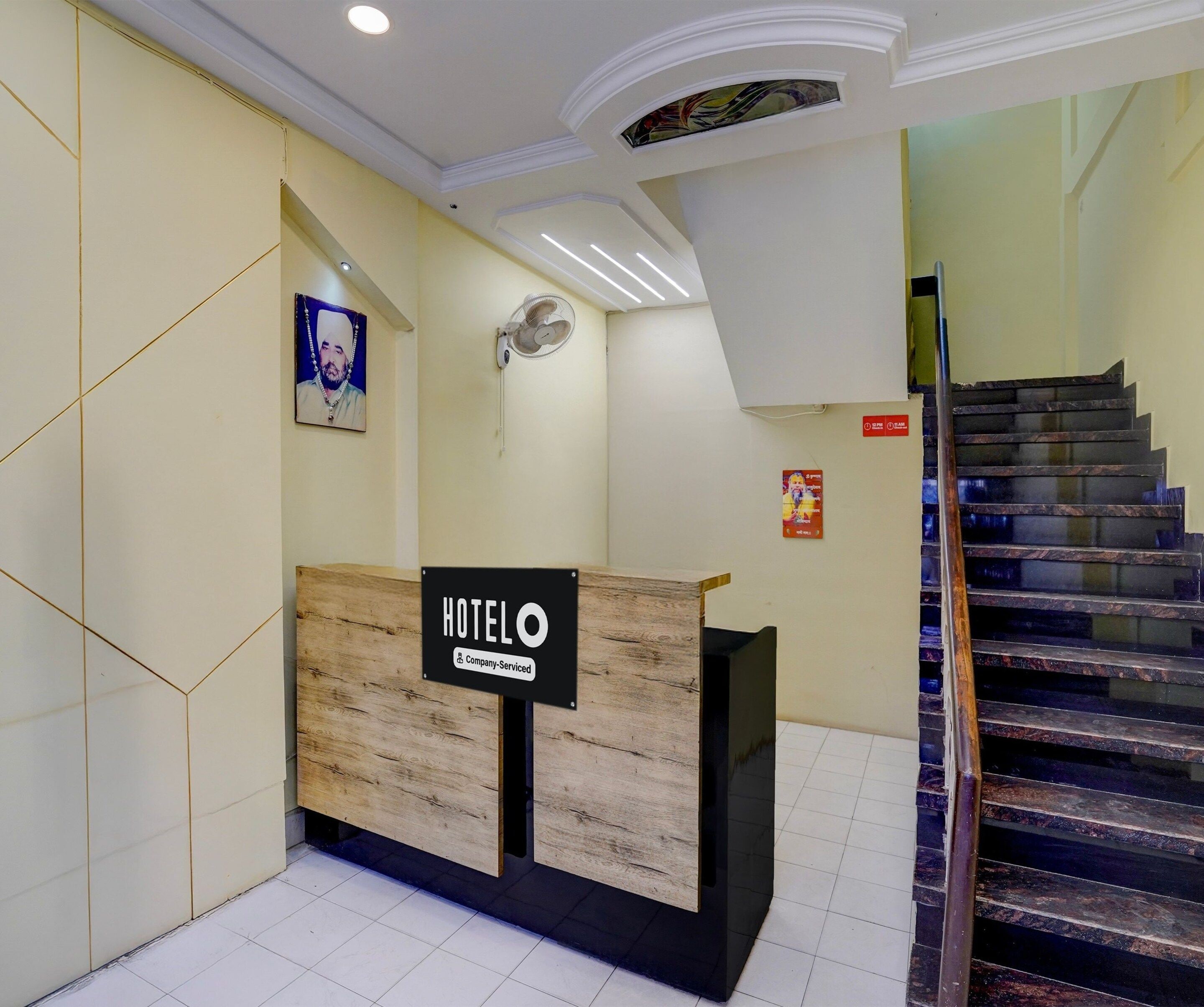 A hotel reception desk with a sign indicating it is company-serviced. There is a staircase to the right and a wall-mounted fan above the desk.