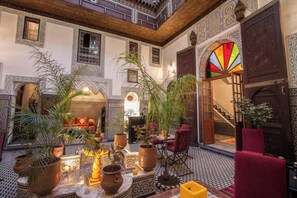 Een traditioneel Marokkaans interieur met versierde tegeltjes, een centrale fontein en een zithoek met een rode bank.