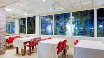 Une salle à manger avec des tables recouvertes de nappes blanches et des chaises rouges, de grandes fenêtres offrant une vue sur la verdure environnante.