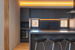 A modern kitchen with dark cabinetry, a central island with stools, and a window with blinds. - Yuki Drift (Kutchan-cho, Abuta-gun)