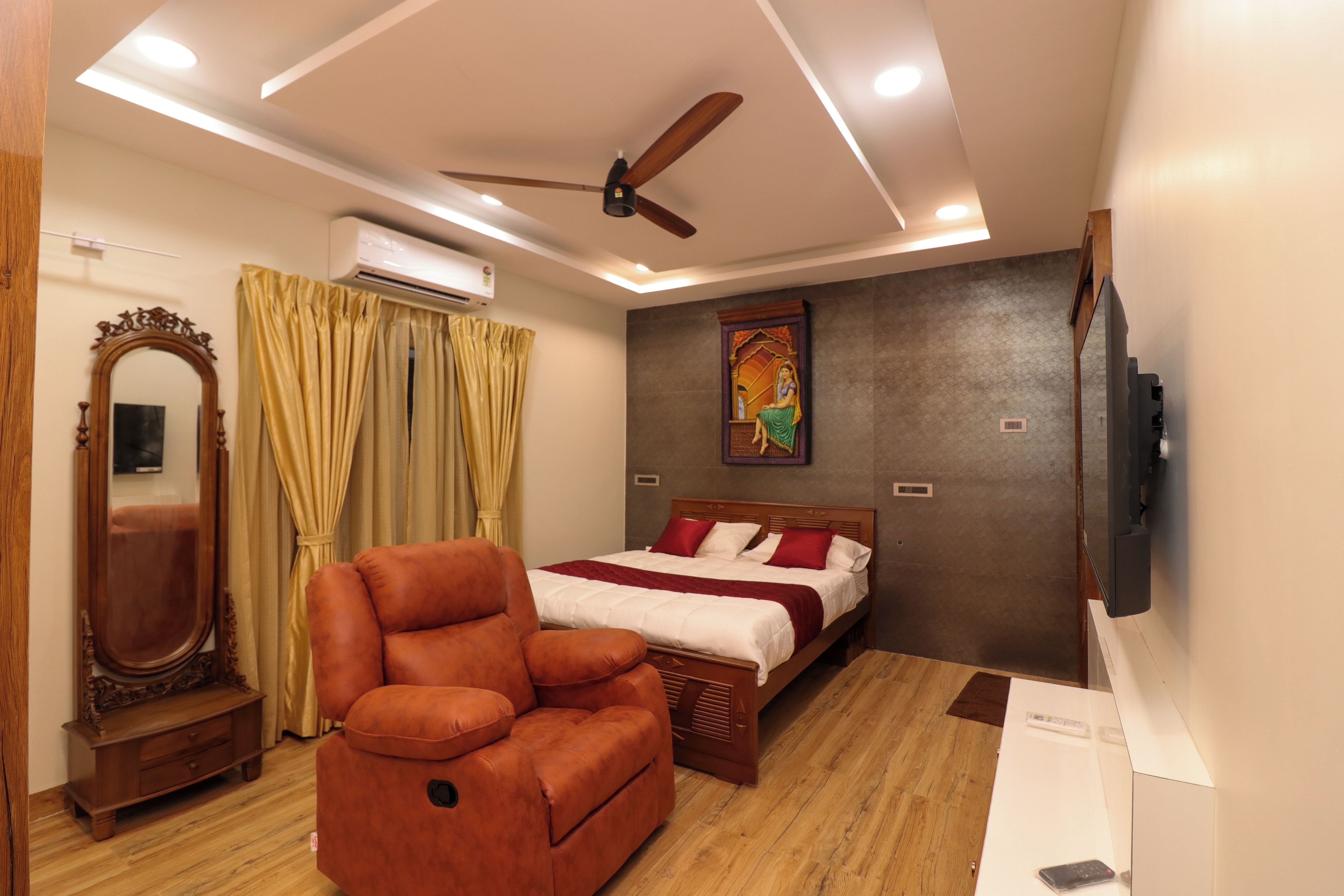 A hotel room with a bed, a sofa, a ceiling fan, an air conditioning unit, a mirror, and a television.