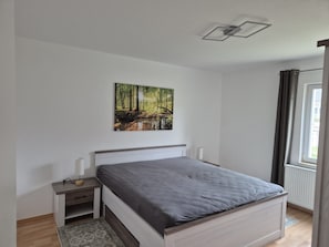 Ein Schlafzimmer mit einem Bett, einem Nachttisch, einer Nachttischlampe, einem Wandbild und einem Fenster mit Vorhängen.