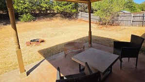 Eine Terrasse im Freien mit Tisch und Stühlen, einem überdachten Bereich und Blick auf einen Garten mit Zaun und Bäumen.