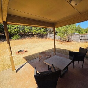 Un patio exterior con mesa y sillas, un espacio cubierto y vista a un jardín trasero con cerco y árboles.