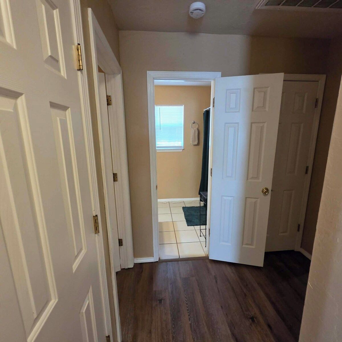 Un couloir avec des portes blanches, une fenêtre visible avec des stores et un sol carrelé.