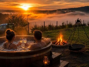 A wooden hot tub with two people, a fire pit, and a view of a sunset over a misty landscape.