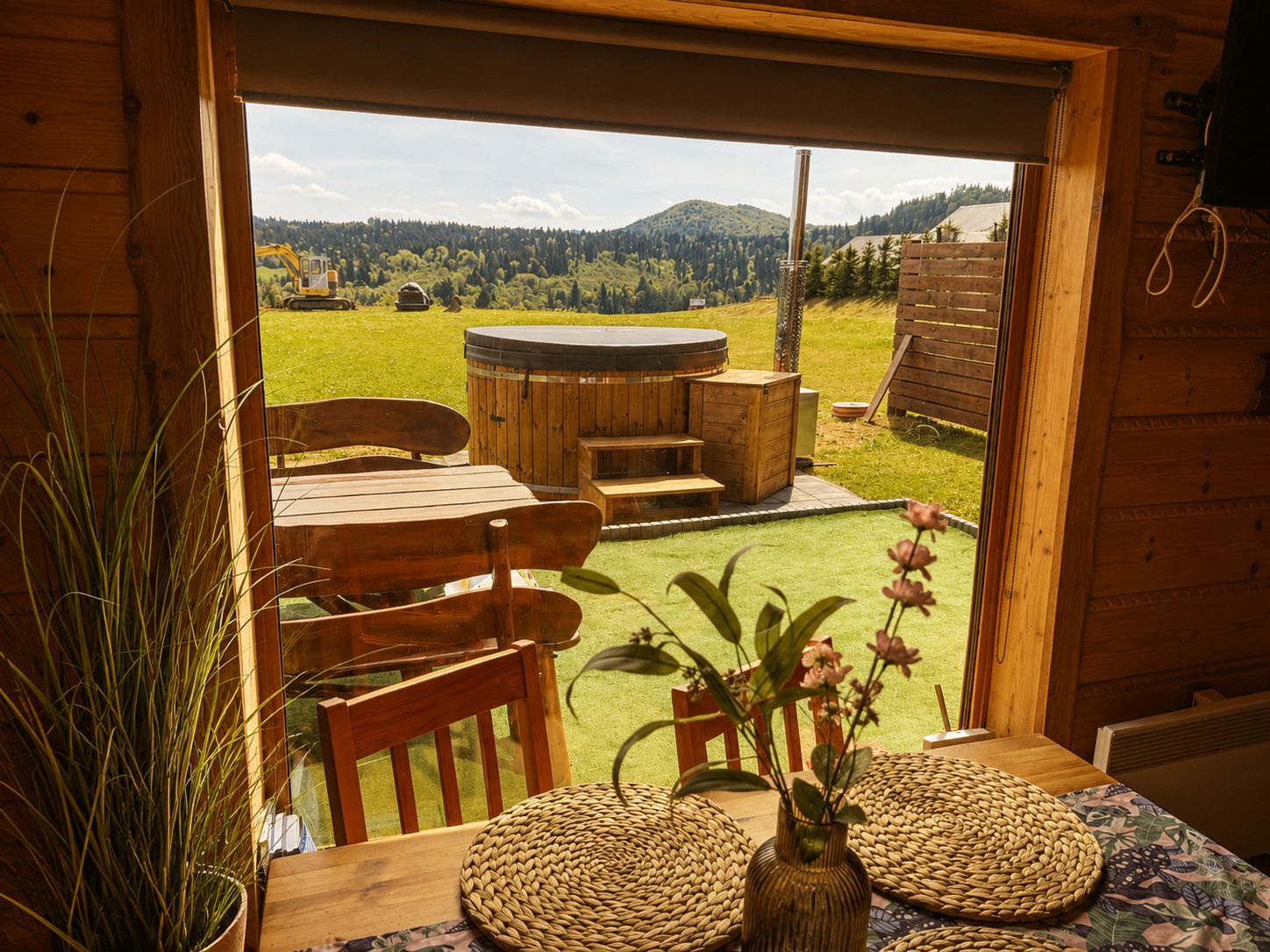 A wooden cabin with a wooden deck, a hot tub, and a view of a green landscape with mountains.
