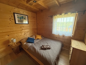 Un dormitorio de cabaña de madera con una cama, almohadas, una mesita de noche, una lámpara y una ventana con cortina.