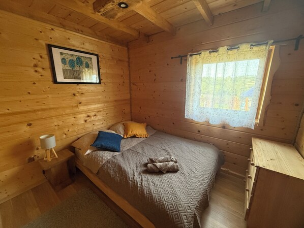 A wooden cabin bedroom with a bed, pillows, a nightstand, a lamp, and a window with a curtain.